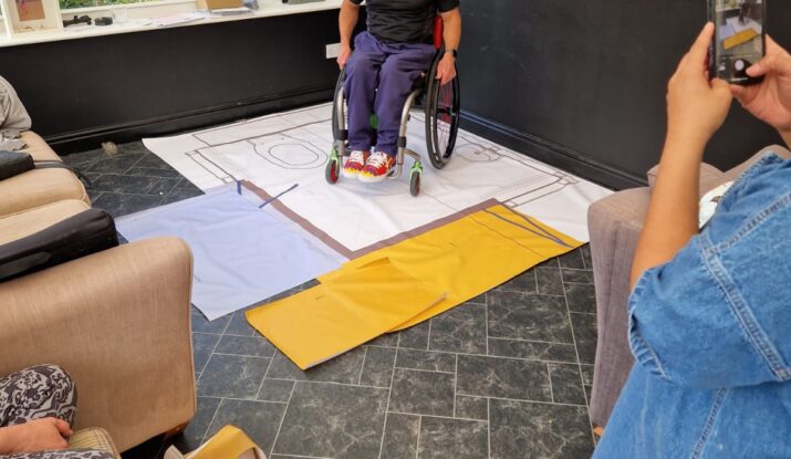 A photo of Ian as he tries out a stencil of a WashPod temporary disabled wetroom for size so that he can imagine how he can manage the space in his wheelchair